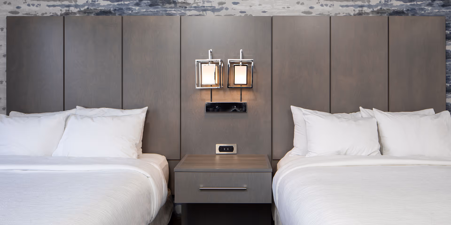 Two neatly made beds with white linens and pillows separated by a wooden nightstand under a modern dual square wall lamp.