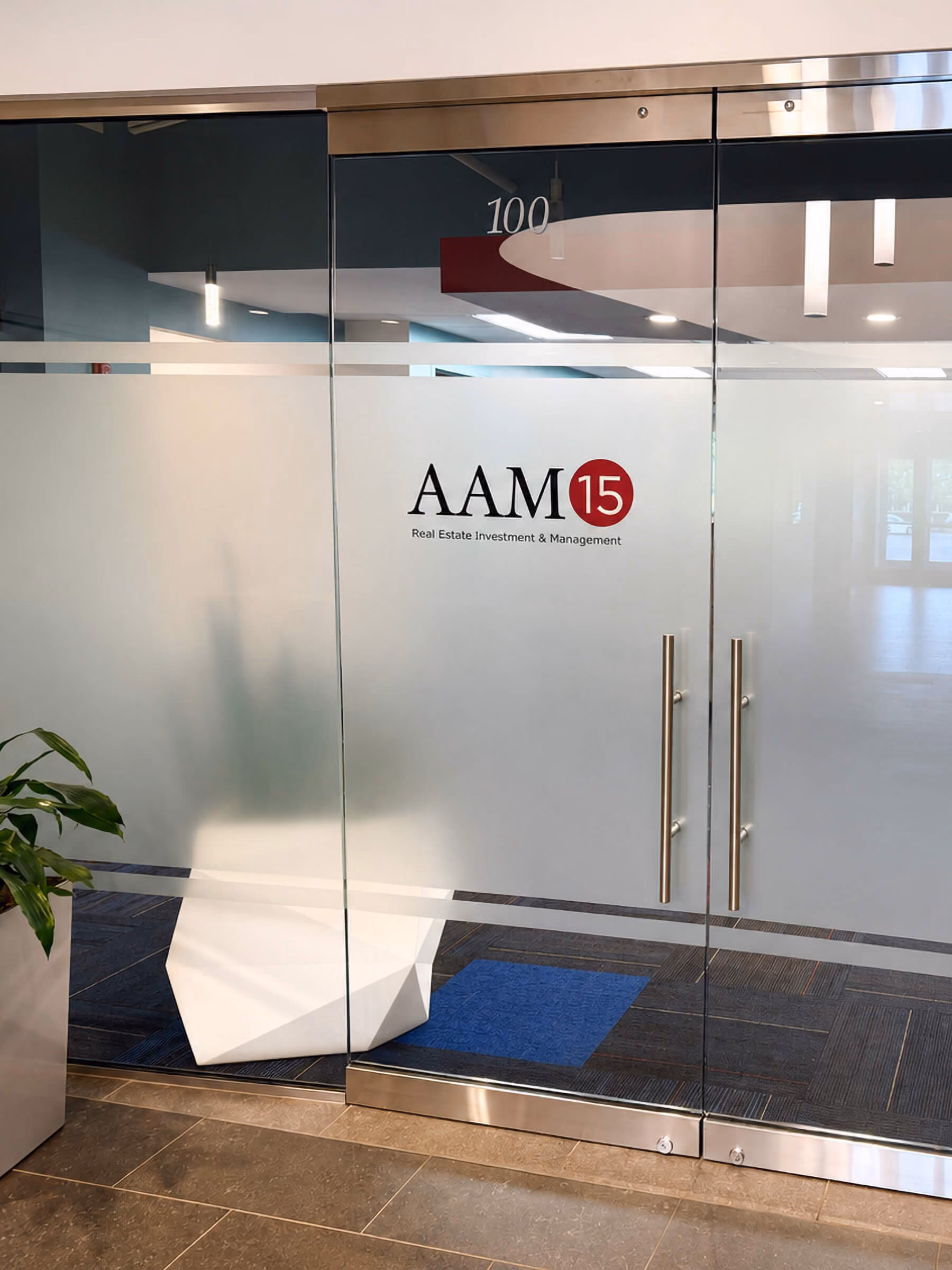 Glass office entrance doors with frosted windows displaying the logo 'AAM 15 Real Estate Investment & Management' and the number 100 above the doors, with a plant on the left and a modern white geometric decorative object inside.