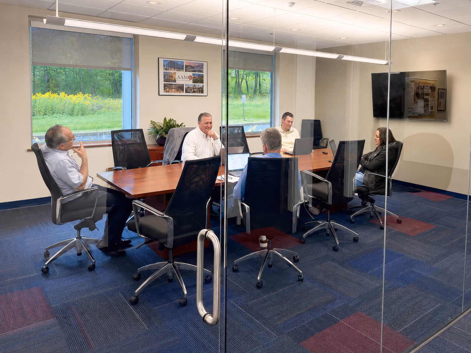 Five people sitting in office chairs around a rectangular wooden conference table in a glass-walled meeting room, with laptops open in front of some participants, two windows showing green trees and yellow flowers outside, a TV mounted on the wall, and a framed collage on the wall.