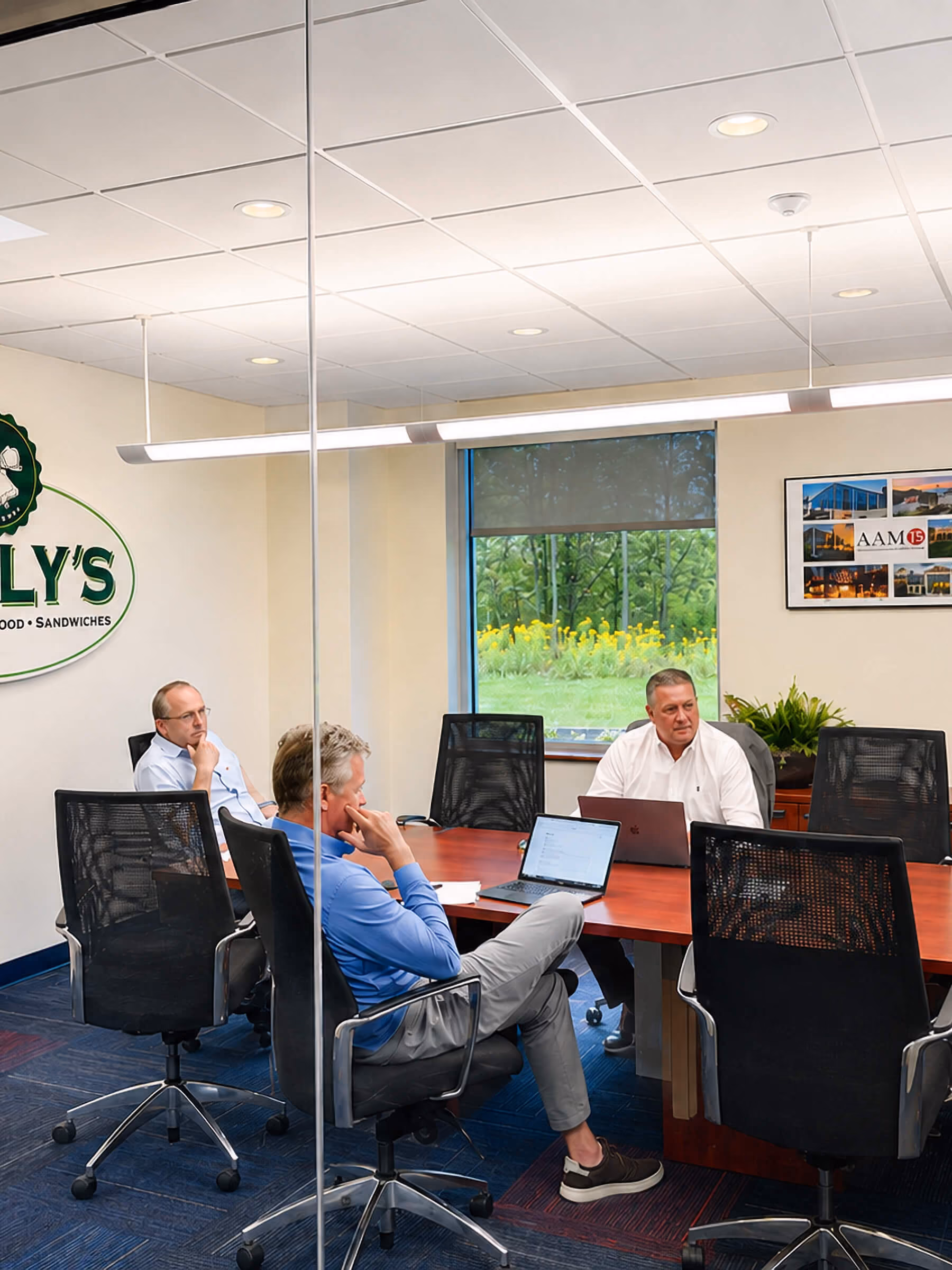 Three men in business casual attire having a meeting in a modern conference room with laptops on a rectangular wooden table, glass walls, and a window showing greenery with yellow flowers outside.