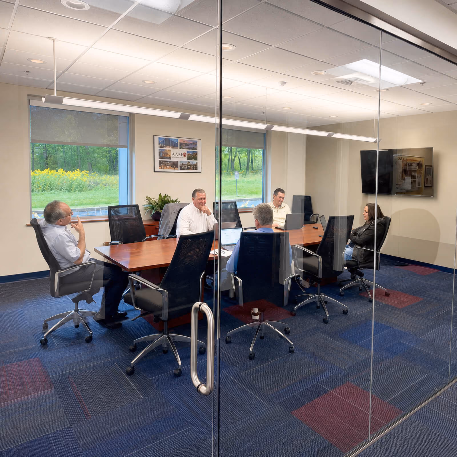 Five people seated around a wooden conference table in a glass-walled office room, with laptops open, two large windows showing green trees and flowers outside, and a wall-mounted TV screen in the background.