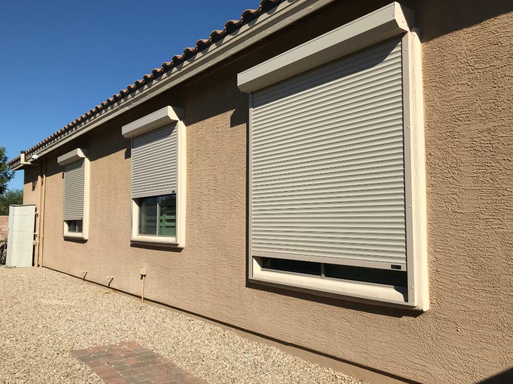 A beige stucco wall of a house with three closed gray roller shutters over the windows. The roof has brown tiles, and there is a small pathway made of bricks leading up to the house. On the left side, there is a white door and a framed area indicating a small outdoor space. The sky is clear and blue, suggesting a sunny day.