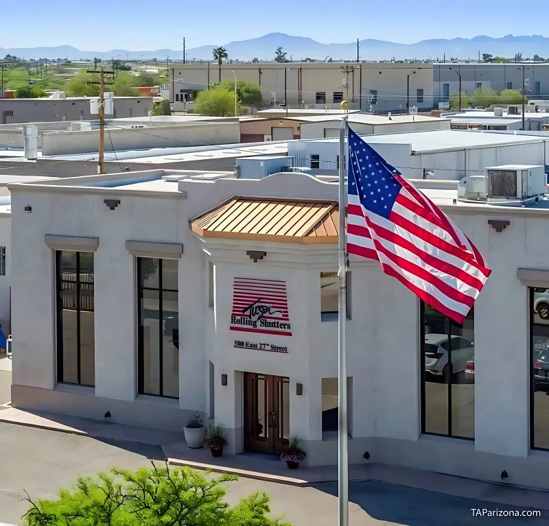 Aerial view of a white stucco building with a distinctive copper-colored roof and large windows. The building features a sign that reads "USA Rolling Shutters" in red and black letters. In front of the building, an American flag is flying on a flagpole. Surrounding the building are various other smaller structures, with greenery visible in the foreground and mountains in the distance under a clear blue sky.