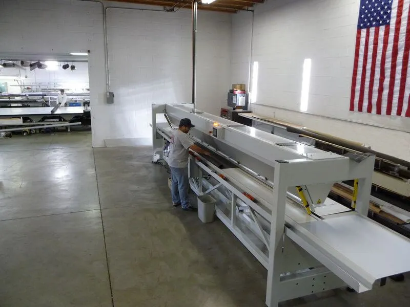 A spacious industrial workspace features a person in a baseball cap working at a large, white machine. The machine has multiple sections and appears to be used for processing materials. A flag hangs on the wall in the background, and there are shelves with items and another worker in the distance, indicating an active working environment. The flooring is concrete, and the lighting is bright, enhancing the clarity of the space.