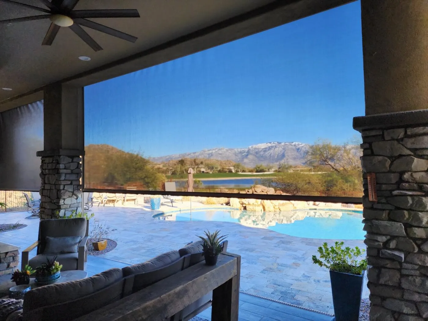 A cozy outdoor patio with a view of a swimming pool and a serene landscape featuring mountains and a body of water. The patio is partially shaded by retractable screens, and there are stone pillars supporting the ceiling. Comfortable seating includes two gray armchairs and a wooden sofa adorned with cushions. A wooden table holds a plant, and there are potted plants in the corners. In the background, a clear blue sky complements the picturesque scenery.