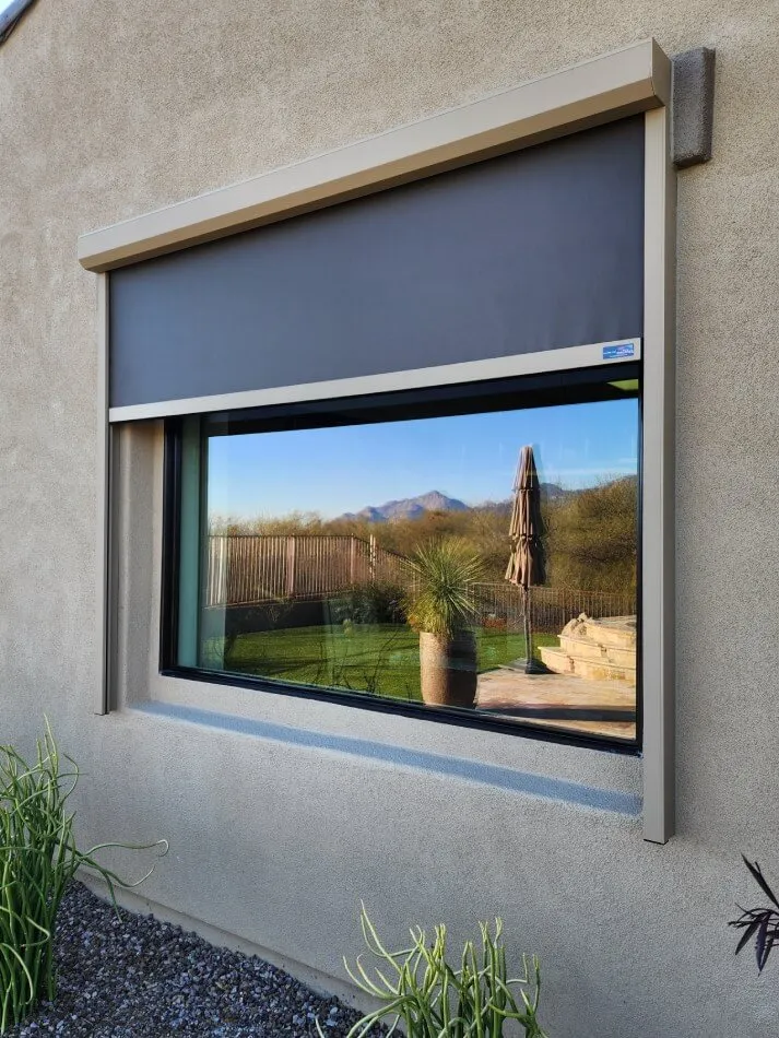 A modern window with dark gray exterior shading is set into a light-colored stucco wall. The window reflects a serene outdoor scene, including green grass and a patio area with a round table and chairs. In the background, there are mountains visible beneath a clear blue sky. An outdoor umbrella stands next to a potted plant on the patio. The foreground features green plants with long leaves growing from gravel.