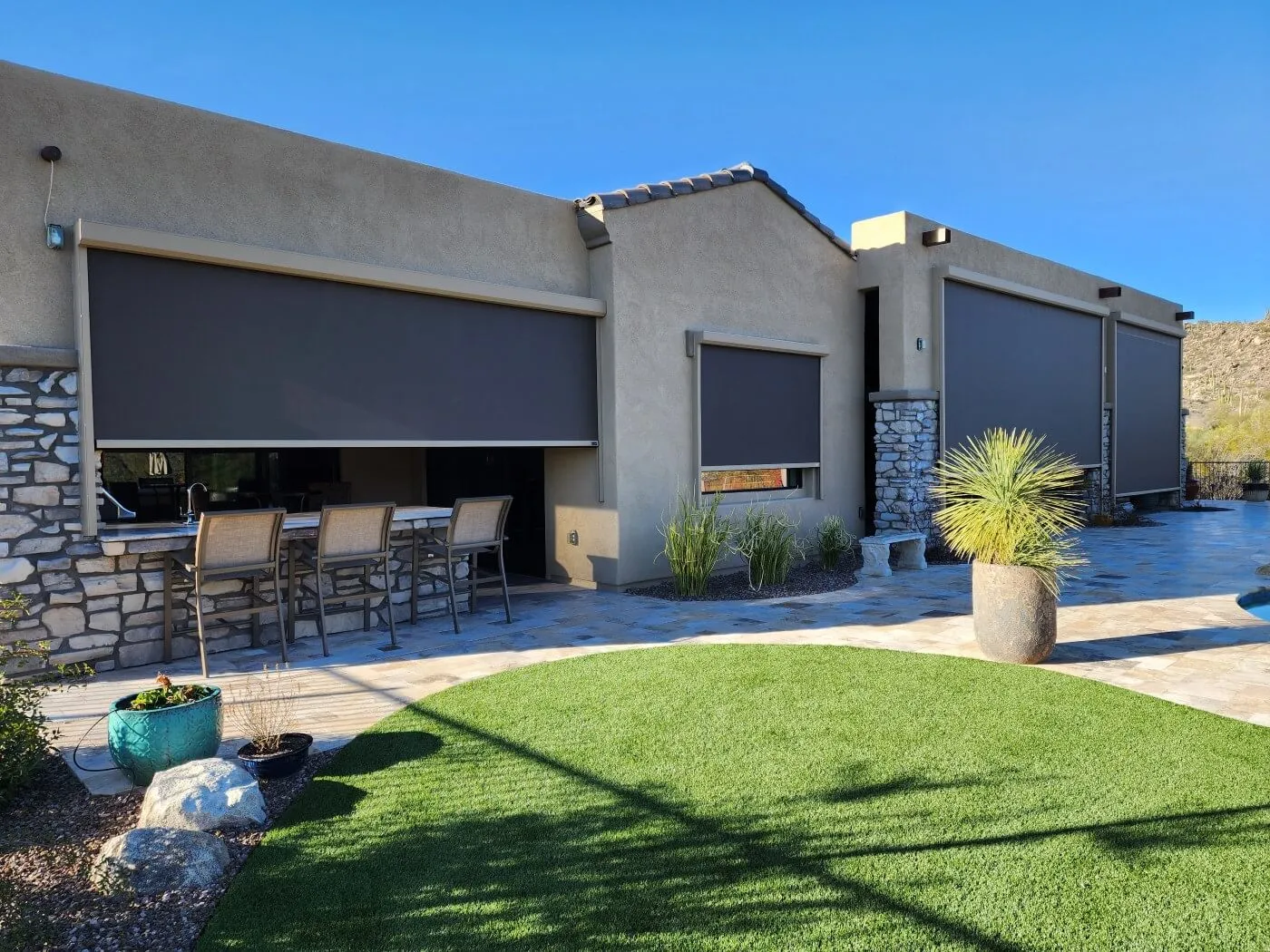 The image shows a modern house exterior with large dark window awnings. The building features a combination of beige stucco and stone elements. In the foreground, there is a small, well-maintained lawn with artificial grass and a circular stone patio. To the left, there is an outdoor bar area with high chairs and a stone countertop. Several potted plants are placed around the patio, contributing to a tidy garden atmosphere. The sky is clear blue, indicating a sunny day.