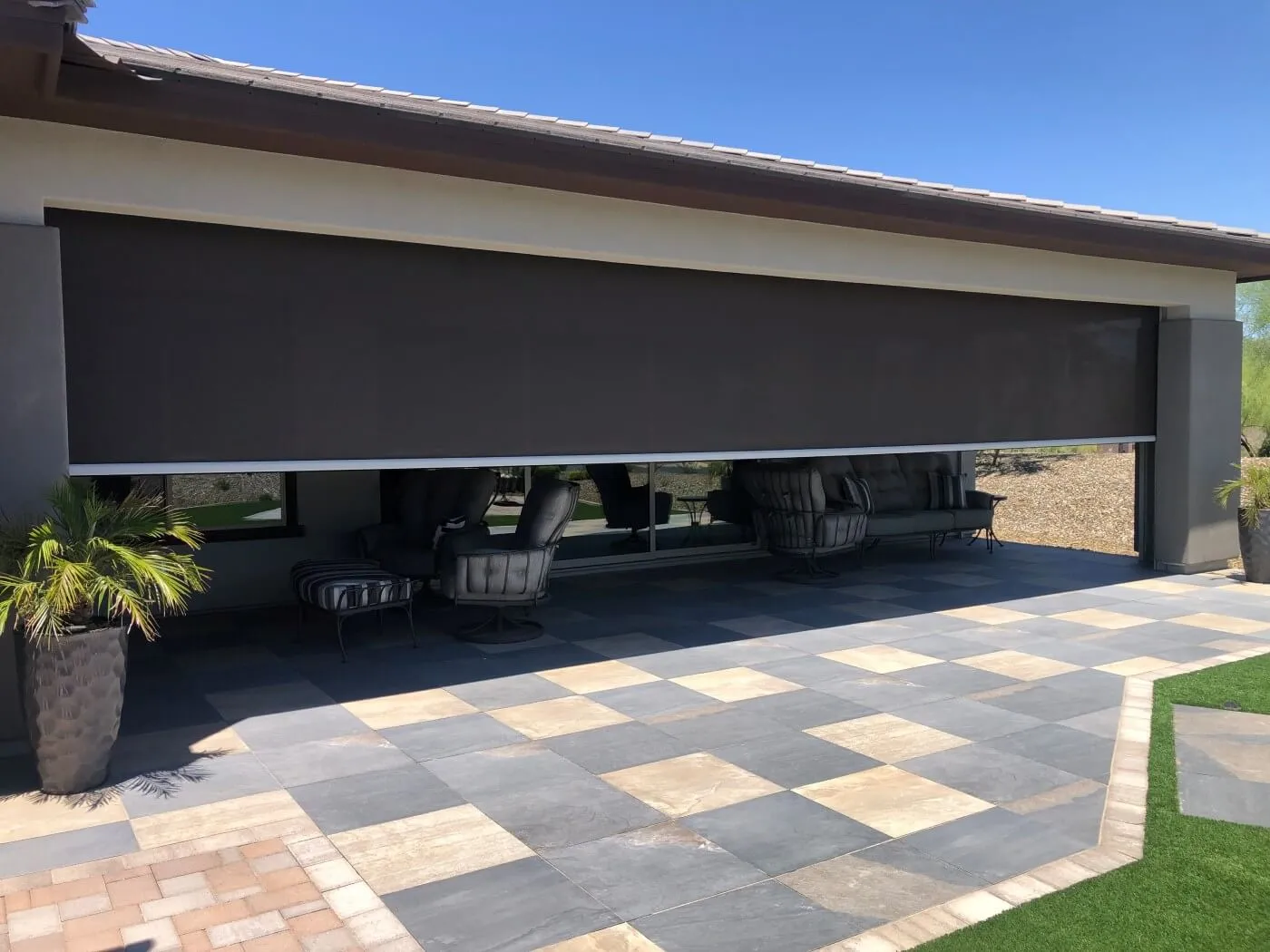 A modern outdoor patio area featuring a large, retractable shade screen covering a window and patio space. The screen is a dark brown color and is fully lowered. Behind it, there are comfortable seating options with gray cushions arranged on a stone-tiled patio. The patio floor has a mix of light and dark stone tiles arranged in a checkerboard pattern. In the foreground, there is a black planter with a green palm plant. The background features a clear blue sky and desert landscape beyond the patio.