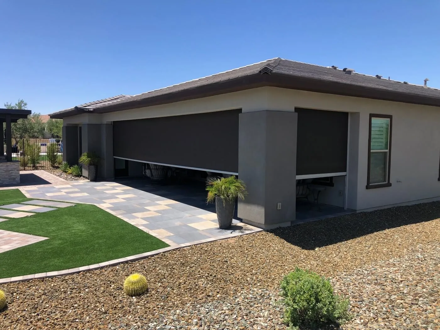 A modern house exterior featuring a garage and a covered patio area. The garage door is equipped with dark outdoor shades that are partially lowered. The patio area is paved with large square tiles and has a section of artificial grass. In the foreground, there are small gravel stones and decorative plants, including a round yellow cactus. The sky is clear and blue, indicating a sunny day.