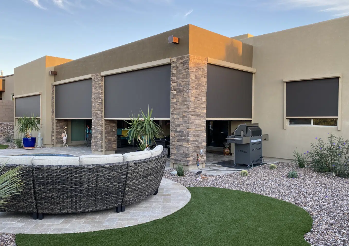 Modern patio with motorized solution screens partially lowered on a beige and stone-clad house, featuring a circular outdoor wicker sofa, artificial turf, gravel landscaping, potted plants, a barbecue grill, and clear evening sky.