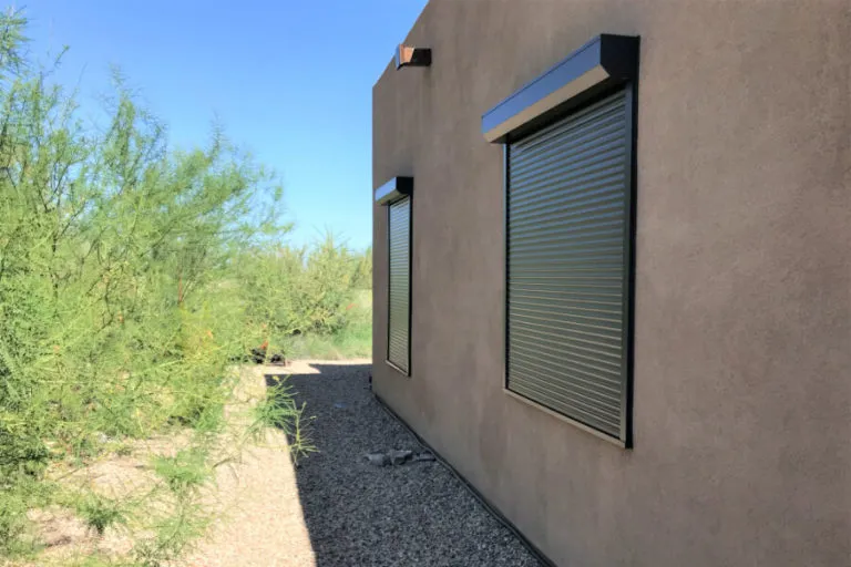 Exterior side view of a stucco building with two windows fully covered by metallic rolling shutters. The surrounding area features desert landscaping with gravel and green shrubs under a clear blue sky.
