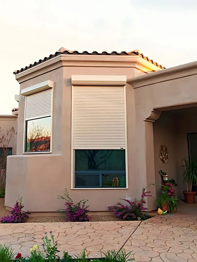 Exterior view of a beige stucco house with a tiled roof, featuring two windows partially covered by white rolling shutters. The house has a landscaped front area with flowering plants and a paved walkway.