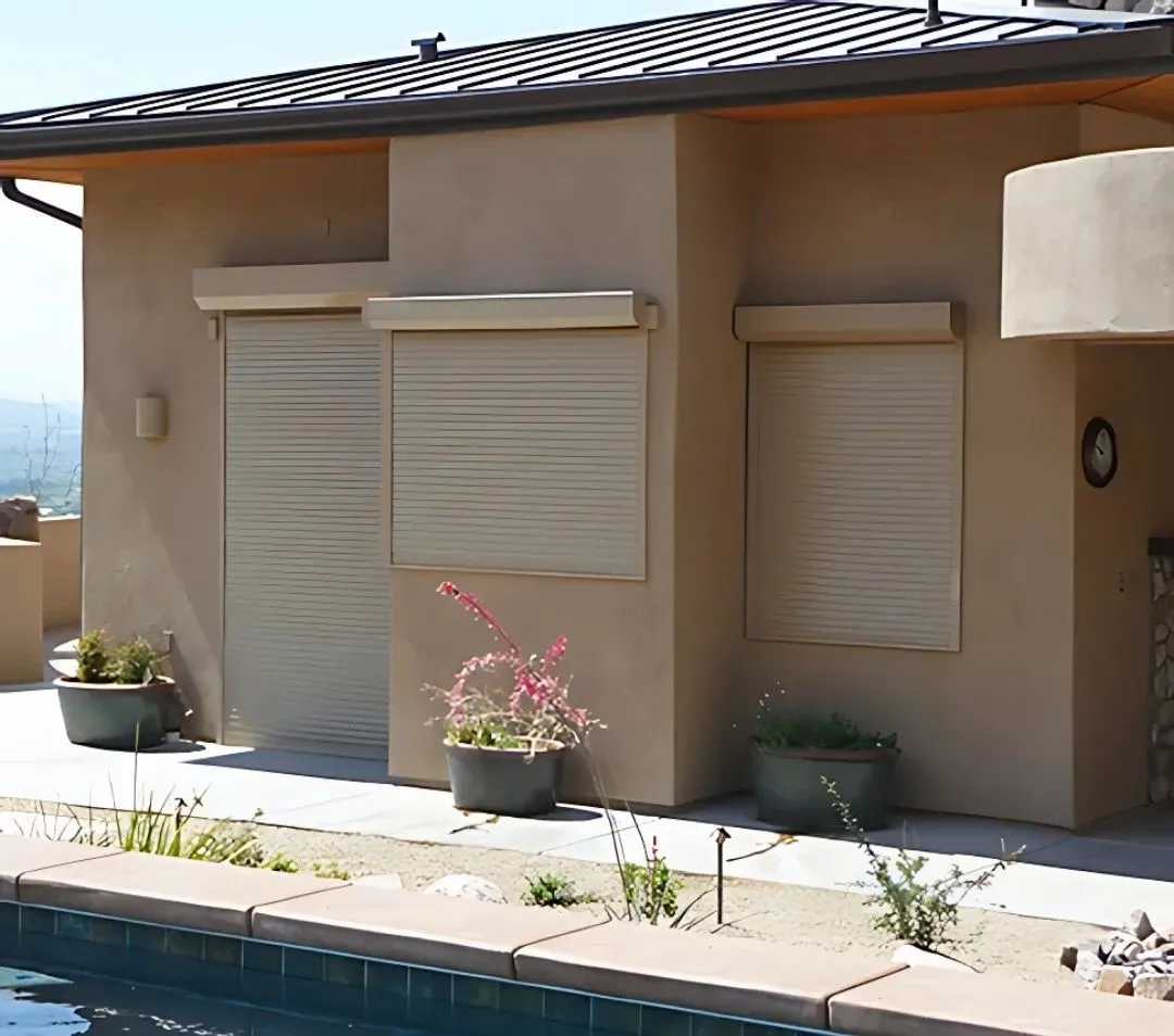 Modern house exterior with three beige rolling shutters fully closed over windows and doors, adjacent to a swimming pool. The stucco walls match the shutters, and potted plants decorate the patio area with desert landscaping.