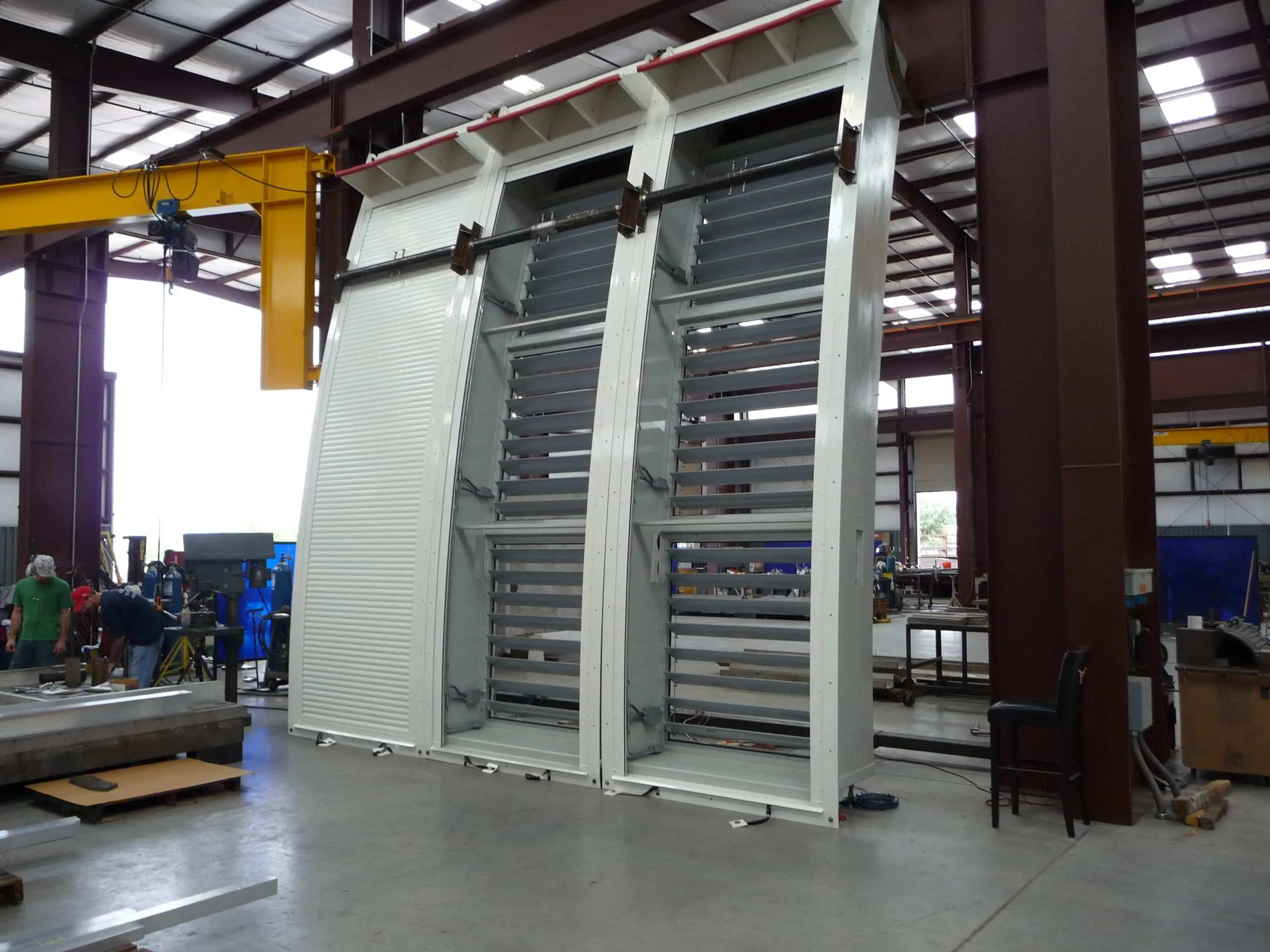 Large industrial metal shutter with adjustable horizontal slats inside a spacious warehouse with workers and machinery in the background.