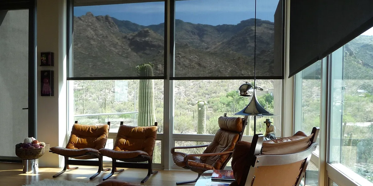 Modern living room with brown leather chairs facing large windows showing a desert landscape with cacti and mountains, some windows partially covered by black roller shades.
