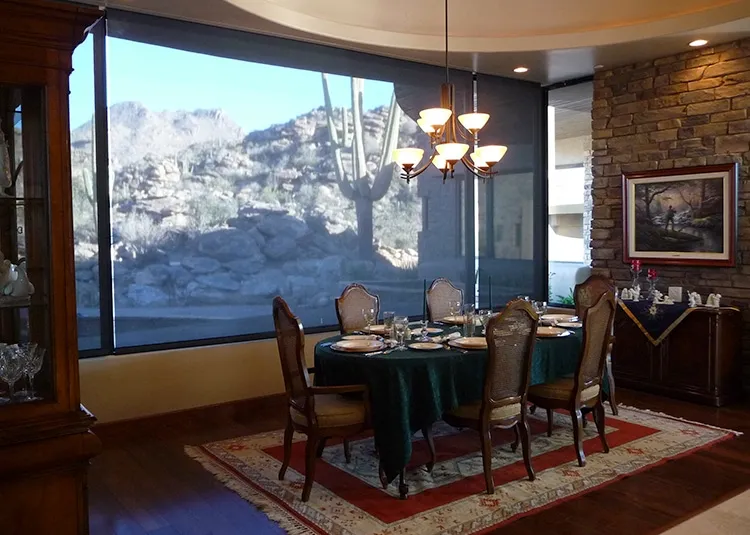 Dining room with a set table for six on a green tablecloth, illuminated by a chandelier and large windows showing rocky desert landscape outside.