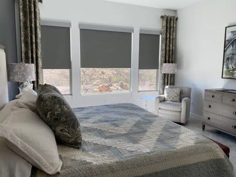 Modern bedroom with a bed featuring patterned bedding, three windows with gray automated blinds partly lowered, two patterned curtains, a white armchair with a cushion, two matching lamps, and a wooden dresser.