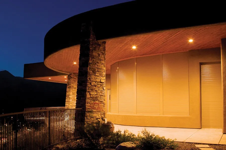 Curved modern house exterior at dusk with stone columns and closed rolling shutters on windows and door.