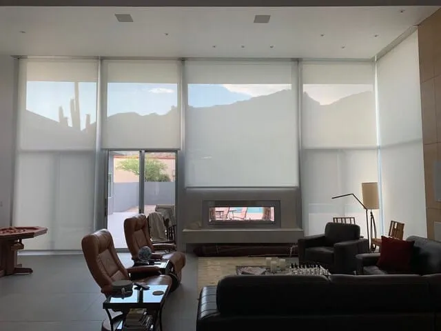 Modern living room with large windows covered by white roller shades, brown leather chairs, a fireplace, and dark sofas.