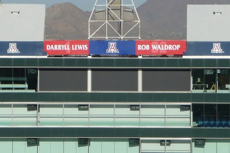 Scoreboard at a stadium displaying names Darryll Lewis and Rob Waldrop with University of Arizona logos, mountains in the background.