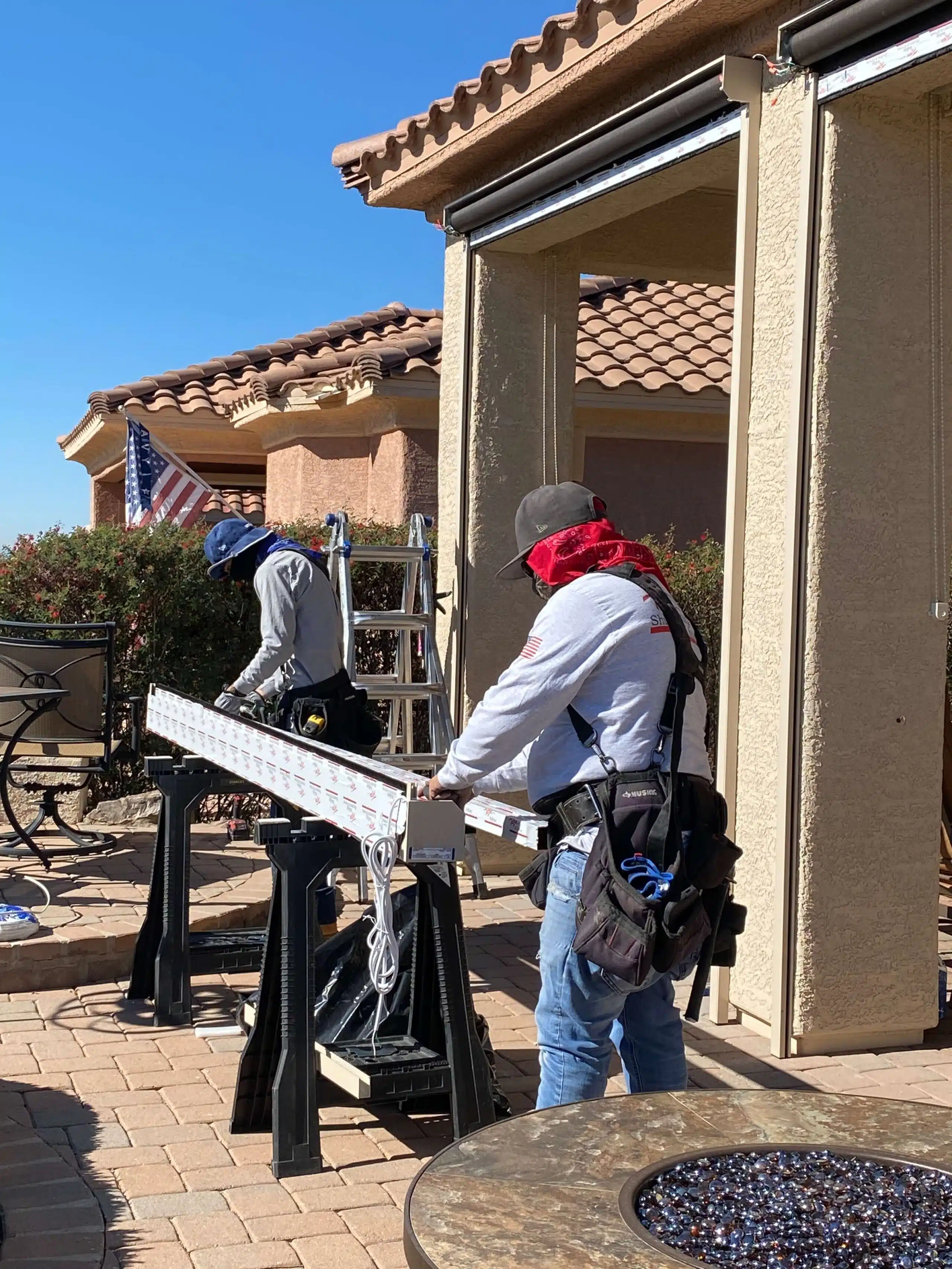 Two workers wearing hats and safety gear working with a long metal piece on sawhorses outside a house with tiled roof under clear blue sky.