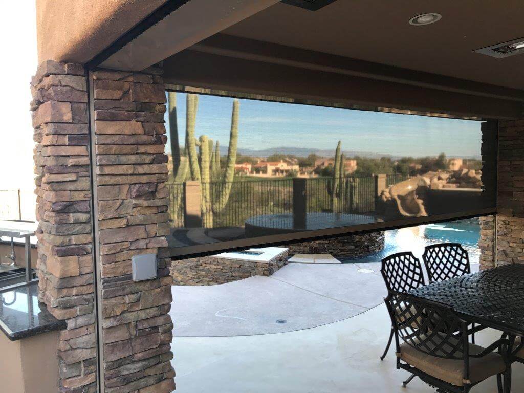 Outdoor patio with stone pillars, black metal dining table and chairs, a pool, and a roll-down screen showing a desert landscape with cacti.