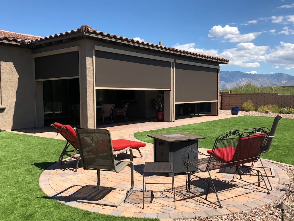 Backyard patio with patio furniture including a red cushioned chaise lounge, red cushioned bench, and metal chairs around a fire pit, with outdoor roller shades on the house and mountains in the background.