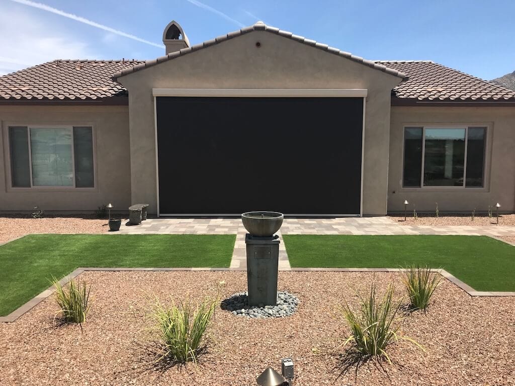Modern single-story house with a large closed black garage door, two windows, and a landscaped front yard featuring gravel, green patches of grass, and a central stone water fountain.