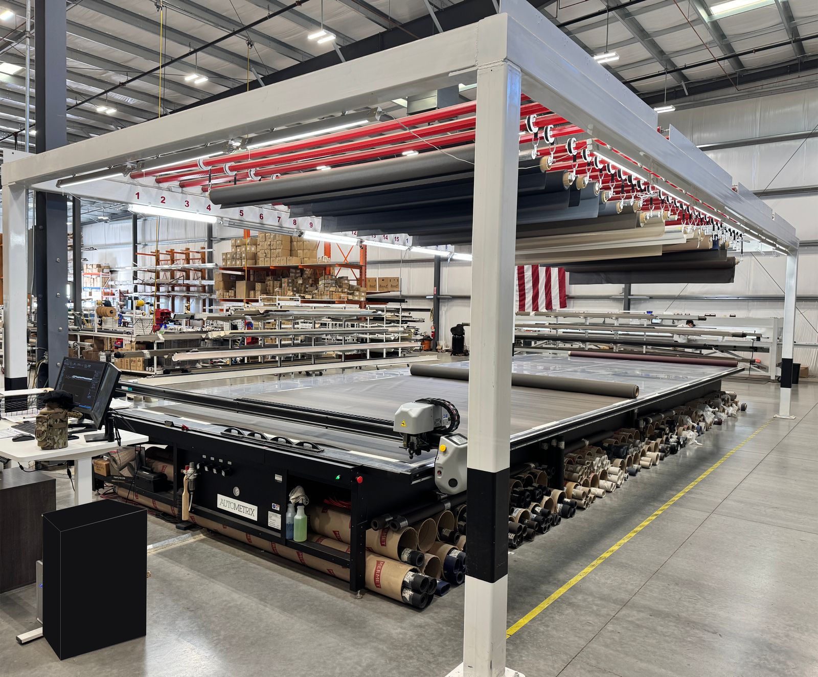 Large automated cutting table in a spacious warehouse with rolls of fabric underneath and an American flag in the background.