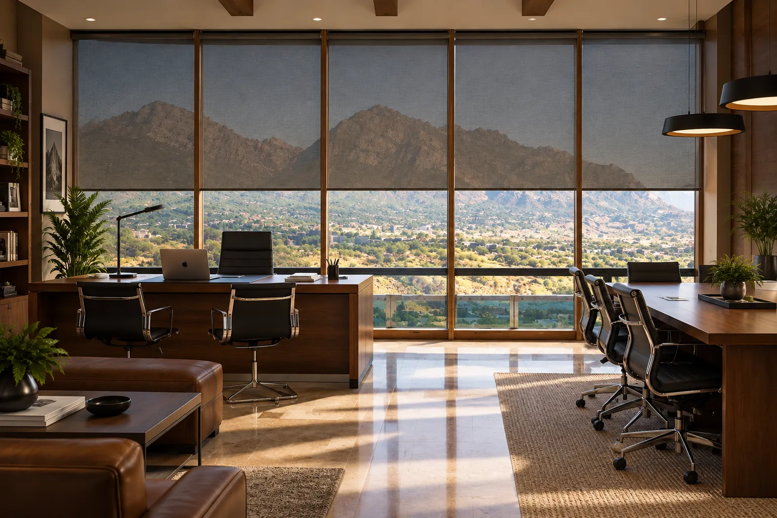 Modern office with large wooden desks and black leather chairs, featuring floor-to-ceiling windows with a view of sunlit mountains and greenery partially covered by gray roller blinds.