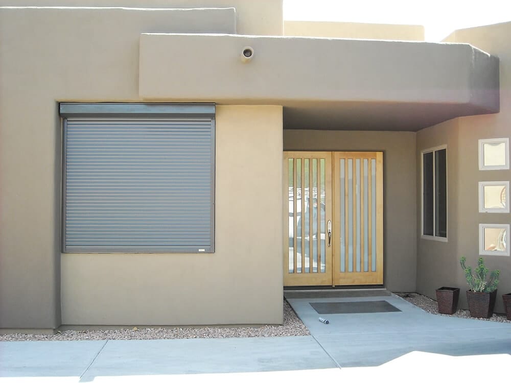 Modern house entrance with closed roller shutter and wooden double doors, surrounded by minimalist landscaping.