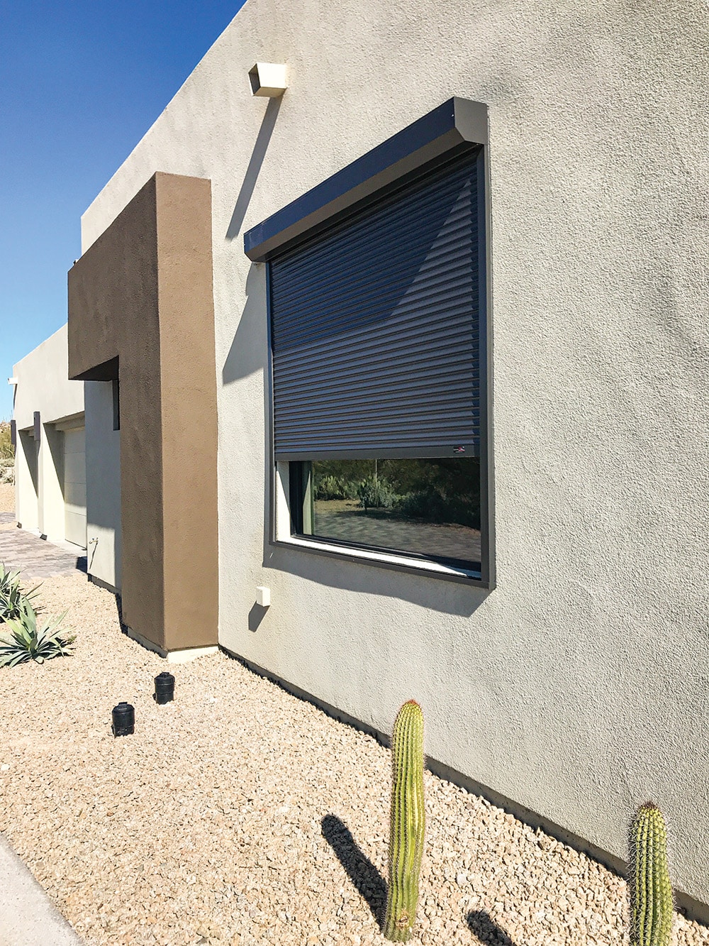 Modern home with a closed dark-colored rolling shutter over a large window, surrounded by a desert landscape with gravel, small cacti, and minimalistic architectural features under a clear blue sky.