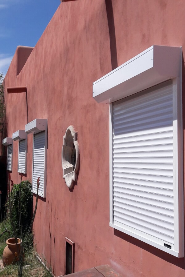 Exterior of a reddish building with multiple closed white rolling shutters over windows, decorative wall ornament, and surrounding greenery under a blue sky.
