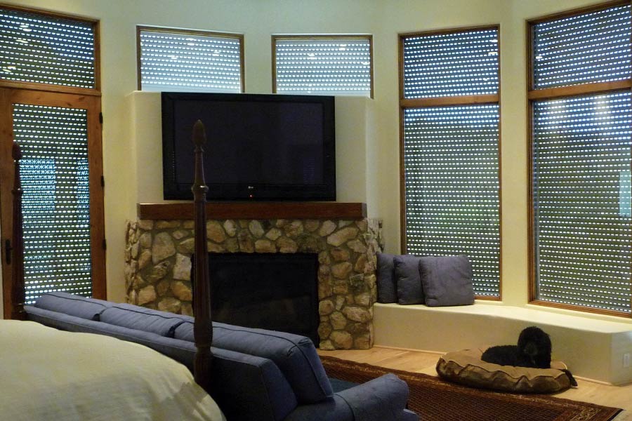 Living room with partially closed rolling shutters on tall windows, allowing filtered light; features include a stone fireplace, wall-mounted TV, cushioned bench seating, and a black dog resting on a round pet bed.