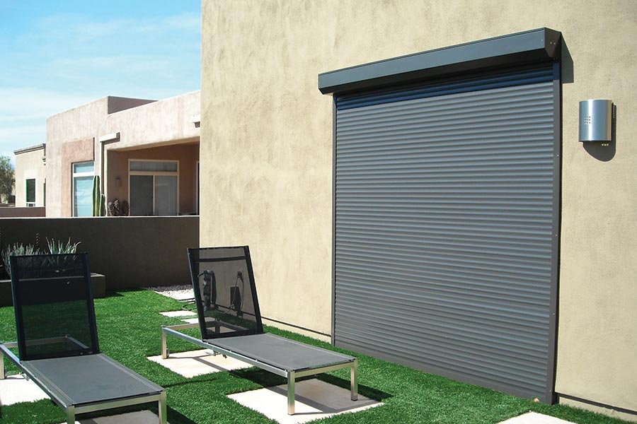 Modern exterior with closed dark gray rolling shutter on a beige stucco wall, artificial grass, two black mesh lounge chairs, and minimalist architecture in a sunny outdoor setting.