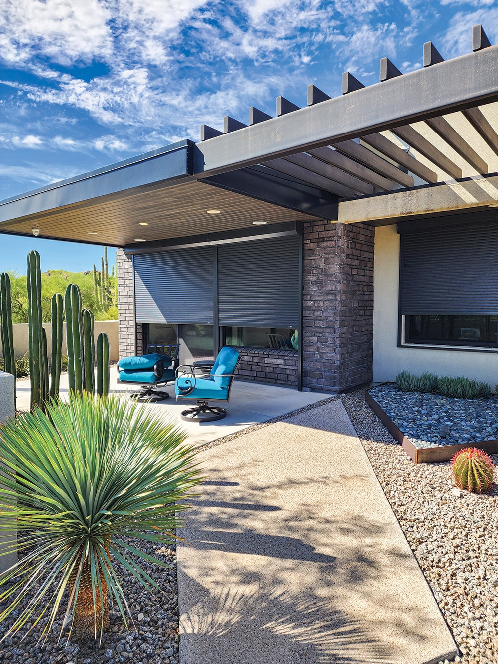 Modern home exterior with rolling shutters partially closed over windows, a shaded patio with two teal cushioned chairs, and a desert landscape featuring cacti, gravel, and a yucca plant under a blue sky with scattered clouds.