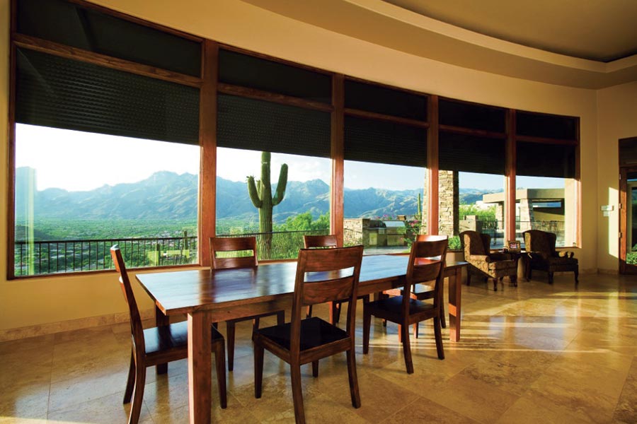 Spacious dining area with large windows partially covered by rolling shutters, offering a panoramic view of desert mountains and a cactus; interior features wooden furniture and polished stone flooring.