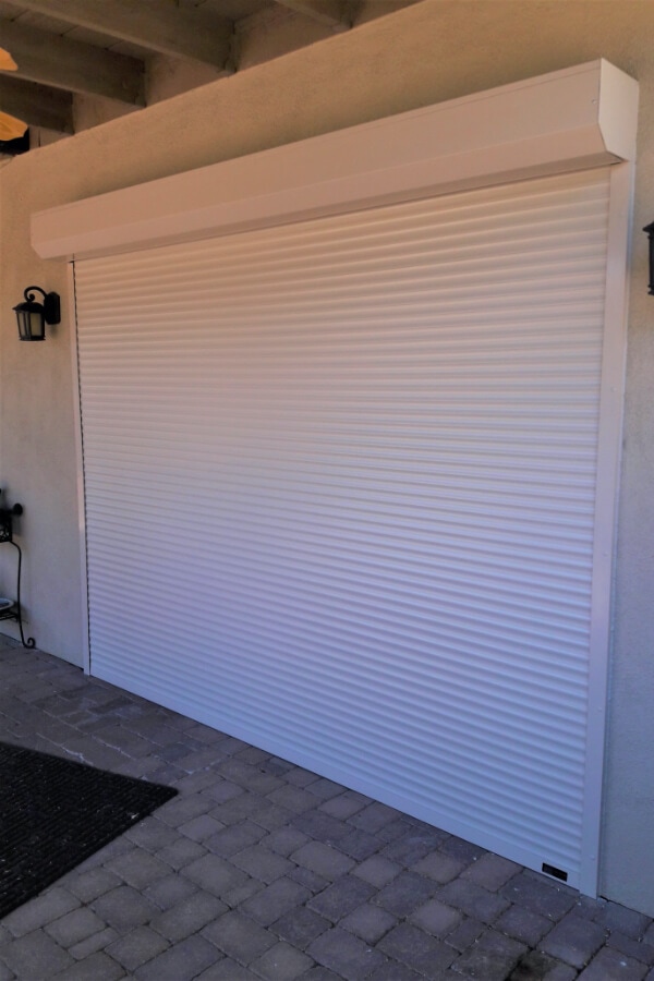 Closed white rolling shutter covering a large opening on an exterior wall, with paver flooring, a doormat, and outdoor wall-mounted lights on either side.