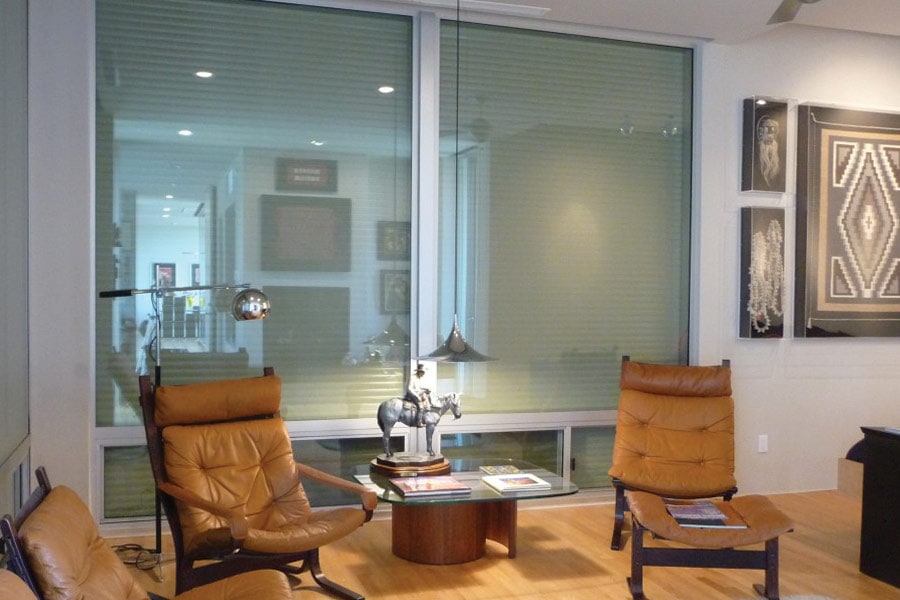 Modern interior space with large windows covered by translucent rolling shutters, two brown leather lounge chairs, a glass-top table with a horse sculpture, wooden flooring, and framed artwork on the wall.
