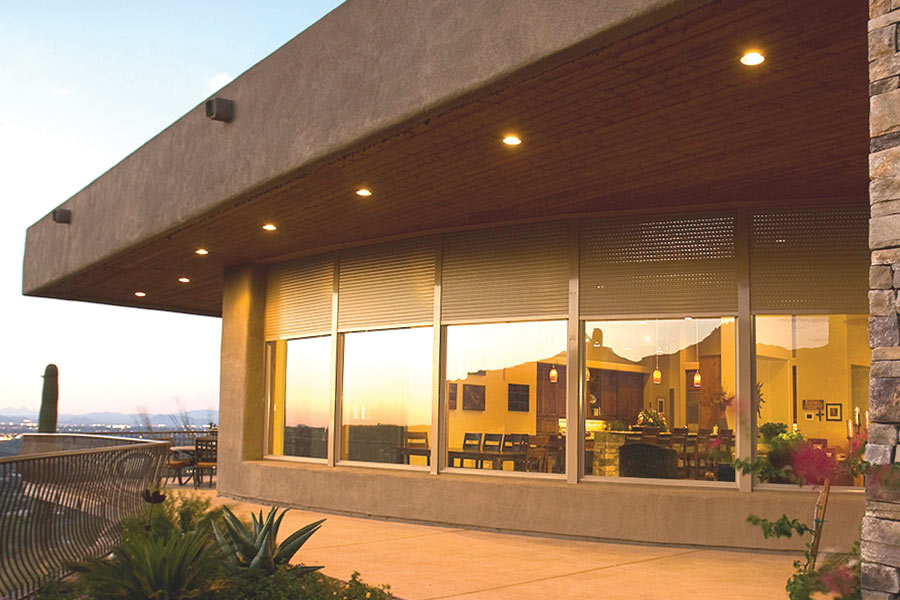 Modern home exterior at sunset with partially open rolling shutters above large curved windows, revealing a well-lit dining and kitchen area inside; features include stone accents, patio lighting, and desert landscaping.
