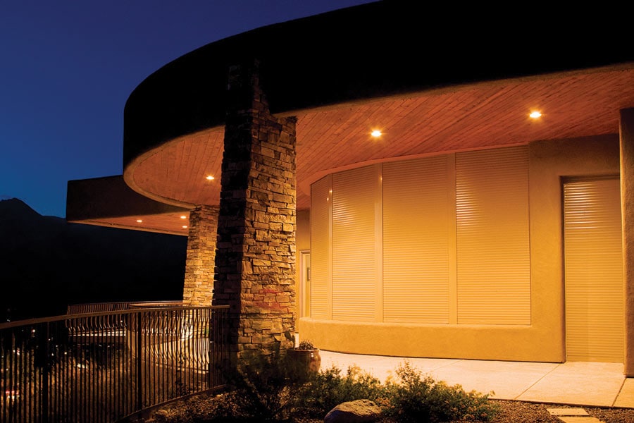 Curved modern home exterior at dusk with warm lighting, featuring closed beige rolling shutters on large windows, a stone pillar, and a metal railing along a landscaped patio.