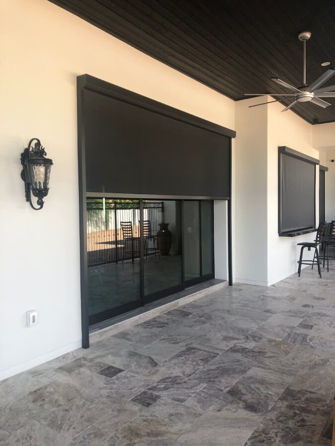 A modern outdoor patio area featuring two large, dark retractable shades that cover glass sliding doors. The shades are mounted on a white wall with a wooden ceiling above. On the left, there is a decorative wall sconce in a vintage style, casting a warm glow. The floor is made of large, patterned tiles in shades of gray and beige. In the background, through the glass doors, some patio furniture is visible along with a potted plant. A ceiling fan is mounted on the ceiling, adding a modern touch to the space.