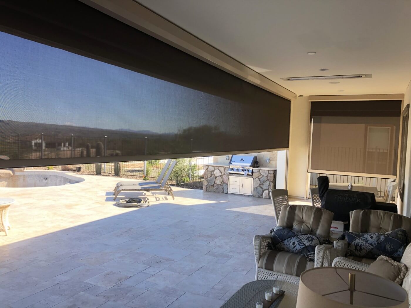 A spacious outdoor patio area is shown with two large window shades partially drawn, allowing some light to filter through. In the foreground, there are two comfortable armchairs with decorative pillows and a small round table beside them. To the left, a pair of sun loungers is positioned near an empty round swimming pool. The background features a built-in outdoor grill and stone counter, surrounded by a fence and landscape, with distant mountains visible in the background under a clear blue sky. The flooring appears to be light-colored stone tiles.