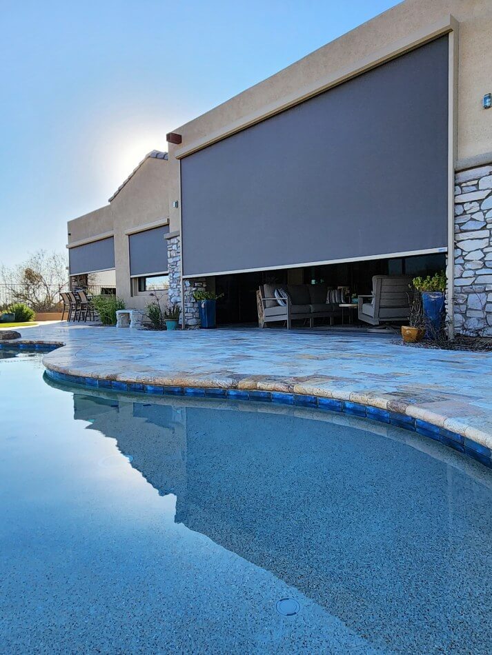 A view of a modern house with large, closed gray outdoor shades on two windows. The house features a stone accent wall and a spacious area with beige patio furniture visible through the shades. In the foreground, there is a circular swimming pool with clear water reflecting the surroundings and a stone border. The sky above is bright and clear, indicating a sunny day. Green plants are placed around the pool area, adding a natural touch to the outdoor setting.