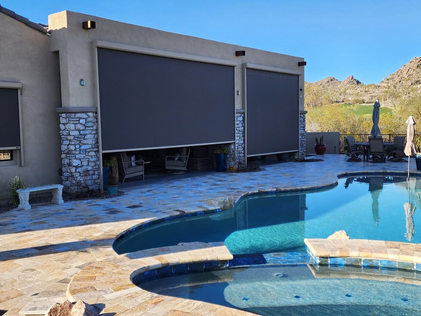 A view of a backyard featuring a swimming pool with a curved edge, surrounded by stone tiles. To the left, there is a small white bench and a planter with green foliage. Two large roll-down shades are installed on a wall of a house, made of a smooth material in a dark gray color, resting over stone accents. In the background, the landscape includes distant mountains and green vegetation under a clear blue sky. There are outdoor chairs visible under one of the shades, and a patio table with umbrellas is situated near the pool.