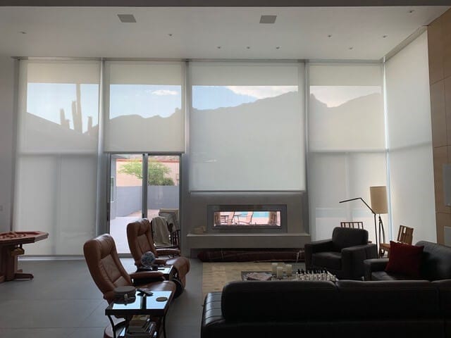 Spacious living room with large floor-to-ceiling windows covered by light-filtering interior shades, modern furniture, a double-sided fireplace, and mountain views partially visible through the shades.