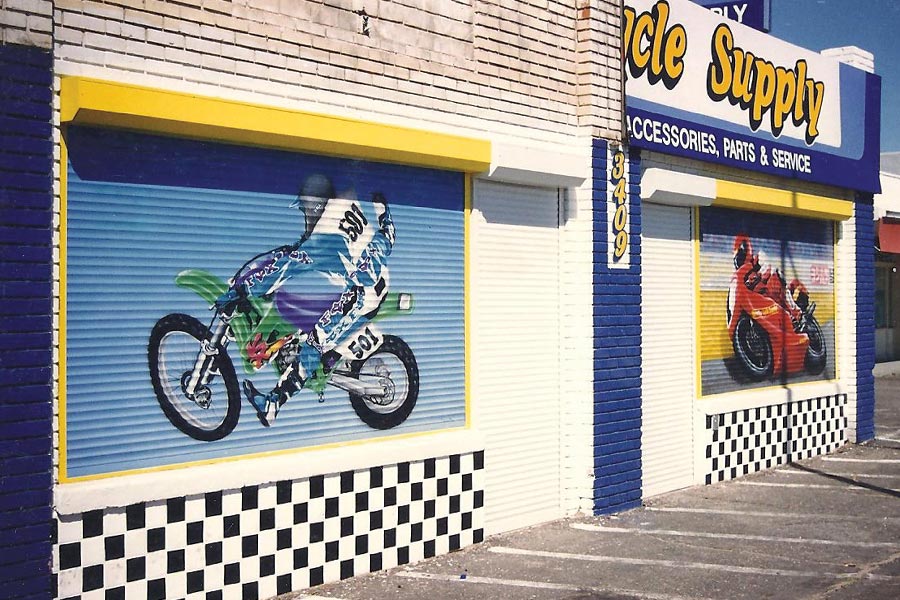 Storefront with two vibrant murals of motorcycles on rolling shutters, yellow and blue signage for a motorcycle supply store, and black and white checkered lower walls