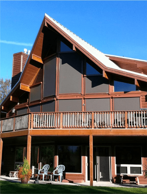 Two-story wooden A-frame building with large triangular and rectangular windows covered by dark solution screens, a wraparound balcony, outdoor seating, and a clear blue sky in the background.