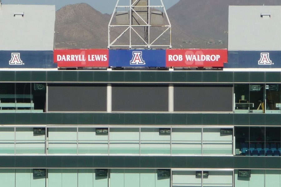 Exterior view of a University of Arizona stadium suite level featuring dark rolling shutters covering some windows, with signage honoring Darryll Lewis and Rob Waldrop, and mountains visible in the background.