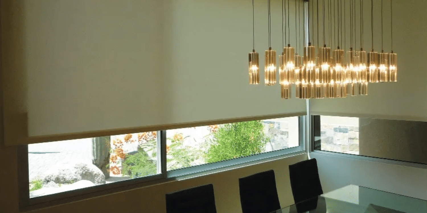 Modern dining area with large windows partially covered by light-colored roller shades; a glass table with dark chairs and a cluster of cylindrical pendant lights hanging above.